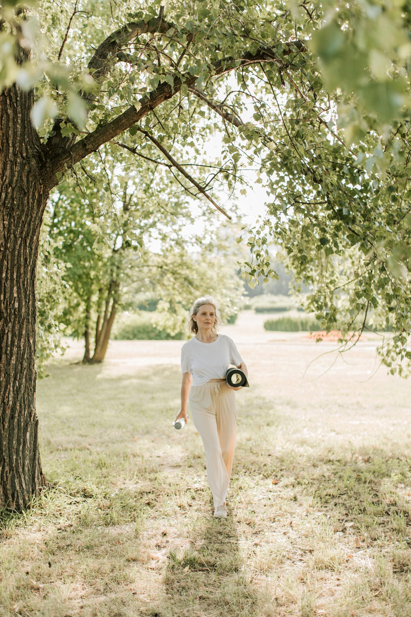 Elderly woman walking with yoga mat in a sunny park, embracing a healthy lifestyle.